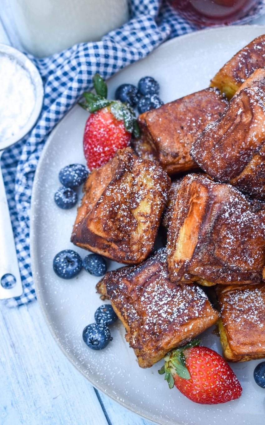 hawaiian roll french toast sprinkled with powdered sugar stacked on a white plate with fresh berries on the side