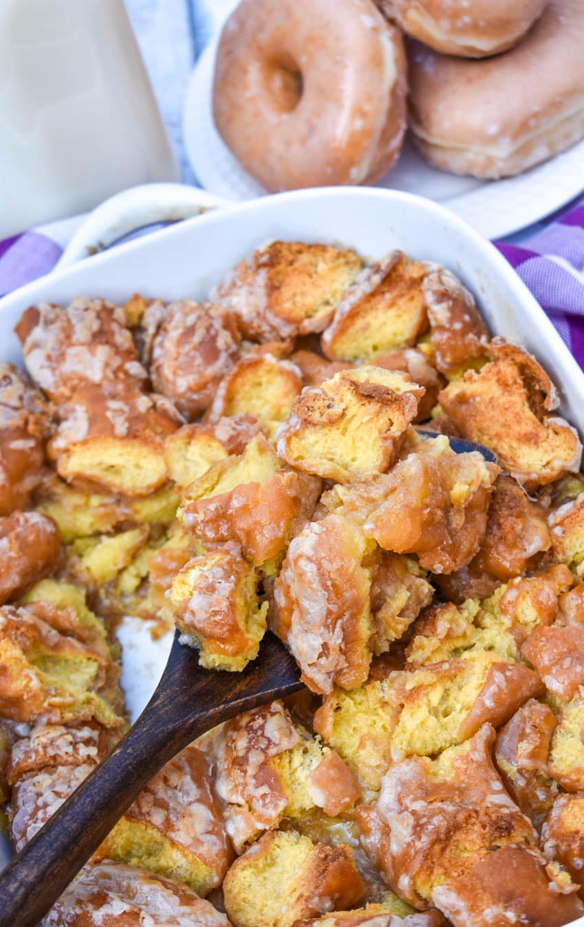 a wooden spatula scooping krispy kreme donut bread pudding out of a white baking dish