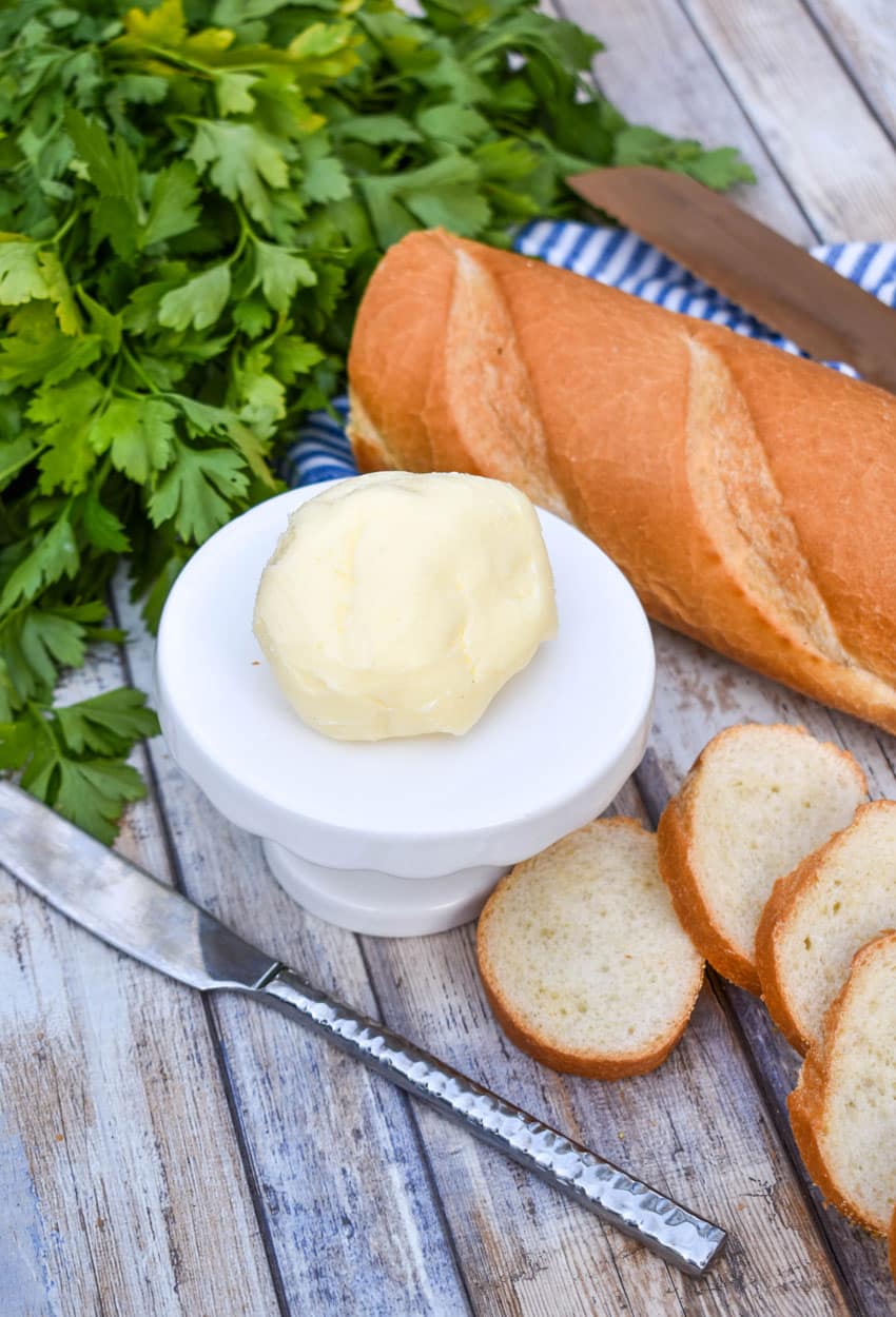 a ball of homemade mason jar butter sitting on a small white dessert stand with slices of baguette bread on the side and fresh herbs in the background
