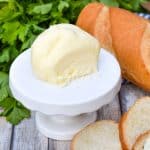 a ball of homemade mason jar butter sitting on a small white dessert stand with slices of baguette bread on the side and fresh herbs in the background