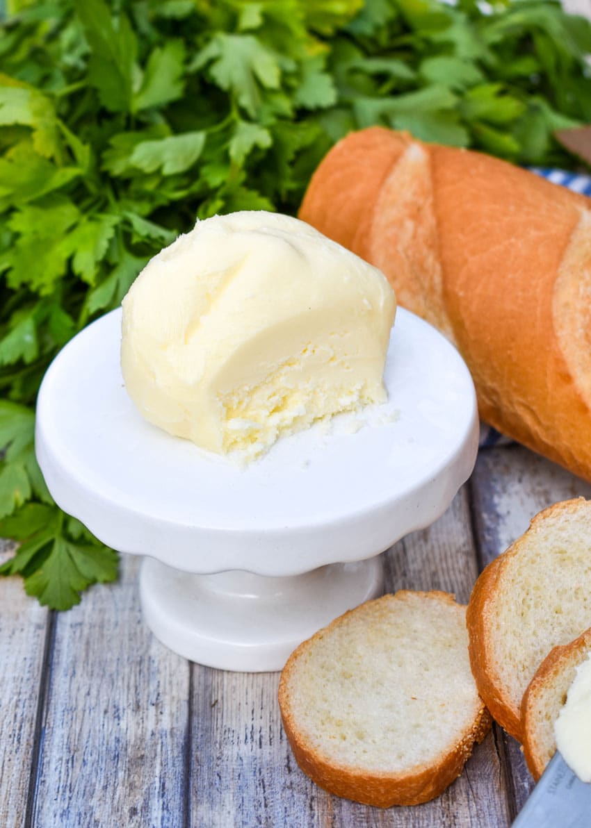 a ball of homemade mason jar butter sitting on a small white dessert stand with slices of baguette bread on the side and fresh herbs in the background