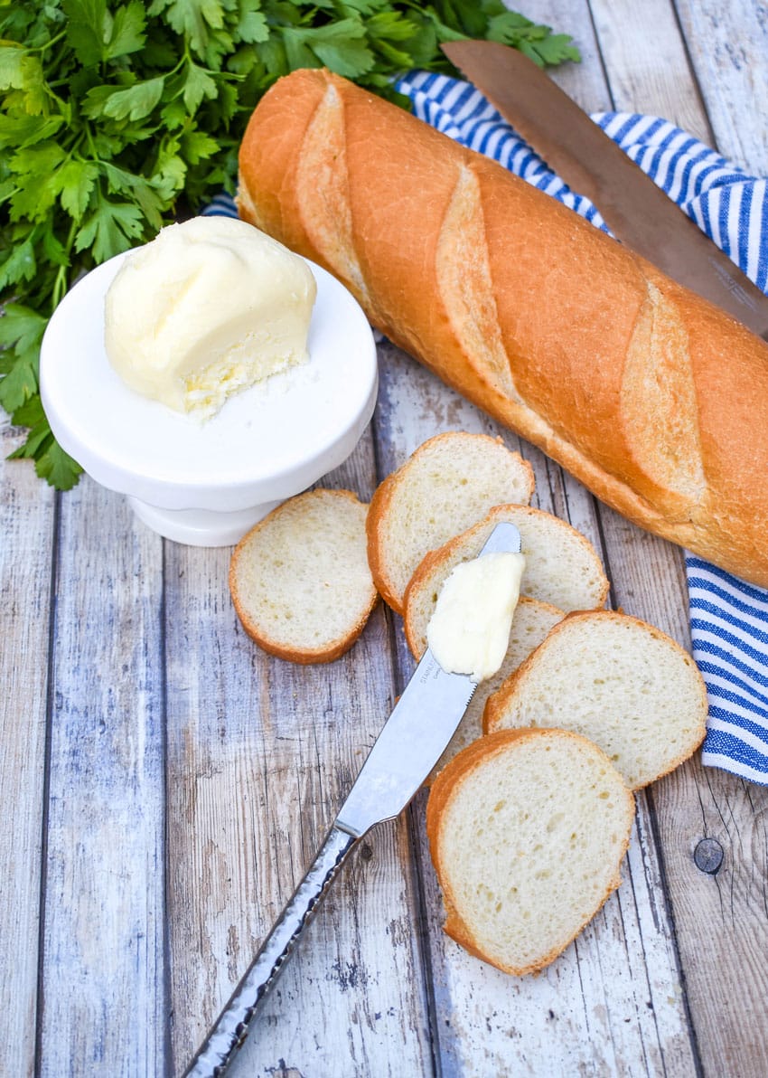 a ball of homemade mason jar butter sitting on a small white dessert stand with slices of baguette bread on the side and fresh herbs in the background