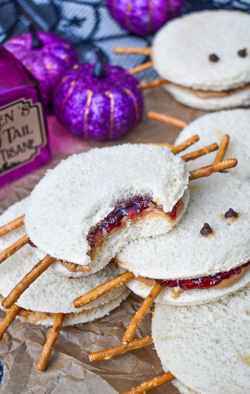 a pile of spider peanut butter and jelly sandwiches on a crinkled sheet of brown parchment paper with a bite taken out of the sandwich on top to reveal the filling inside