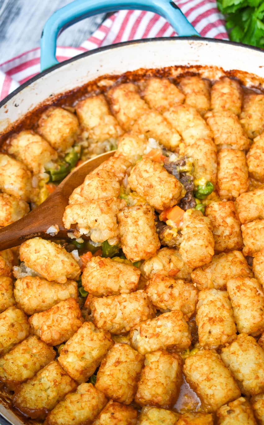 a wooden spoon scooping shepherds pie tater tot casserole out of a blue enameled cast iron skillet
