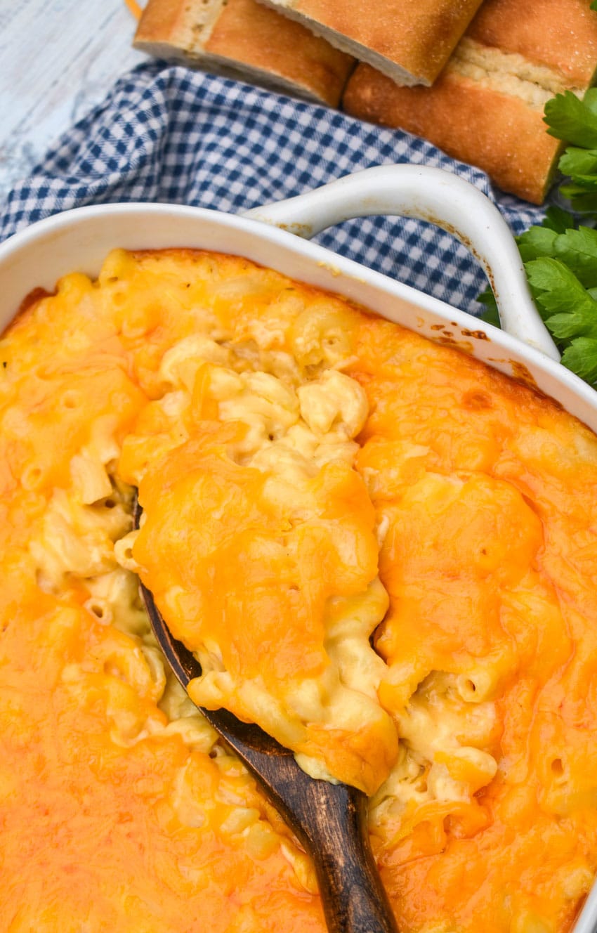 a wooden spoon scooping extra creamy macaroni and cheese out of a white baking dish