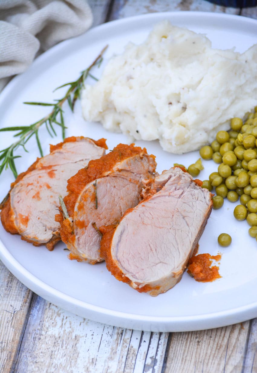 slices of roasted pumpkin butter pork on a white plate with mashed potatoes and peas on the side