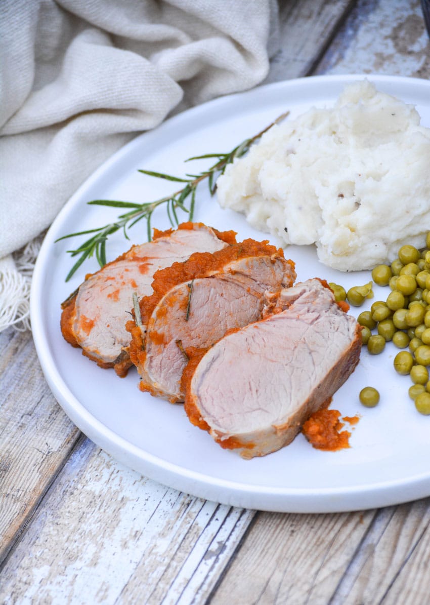 slices of roasted pumpkin butter pork on a white plate with mashed potatoes and peas on the side