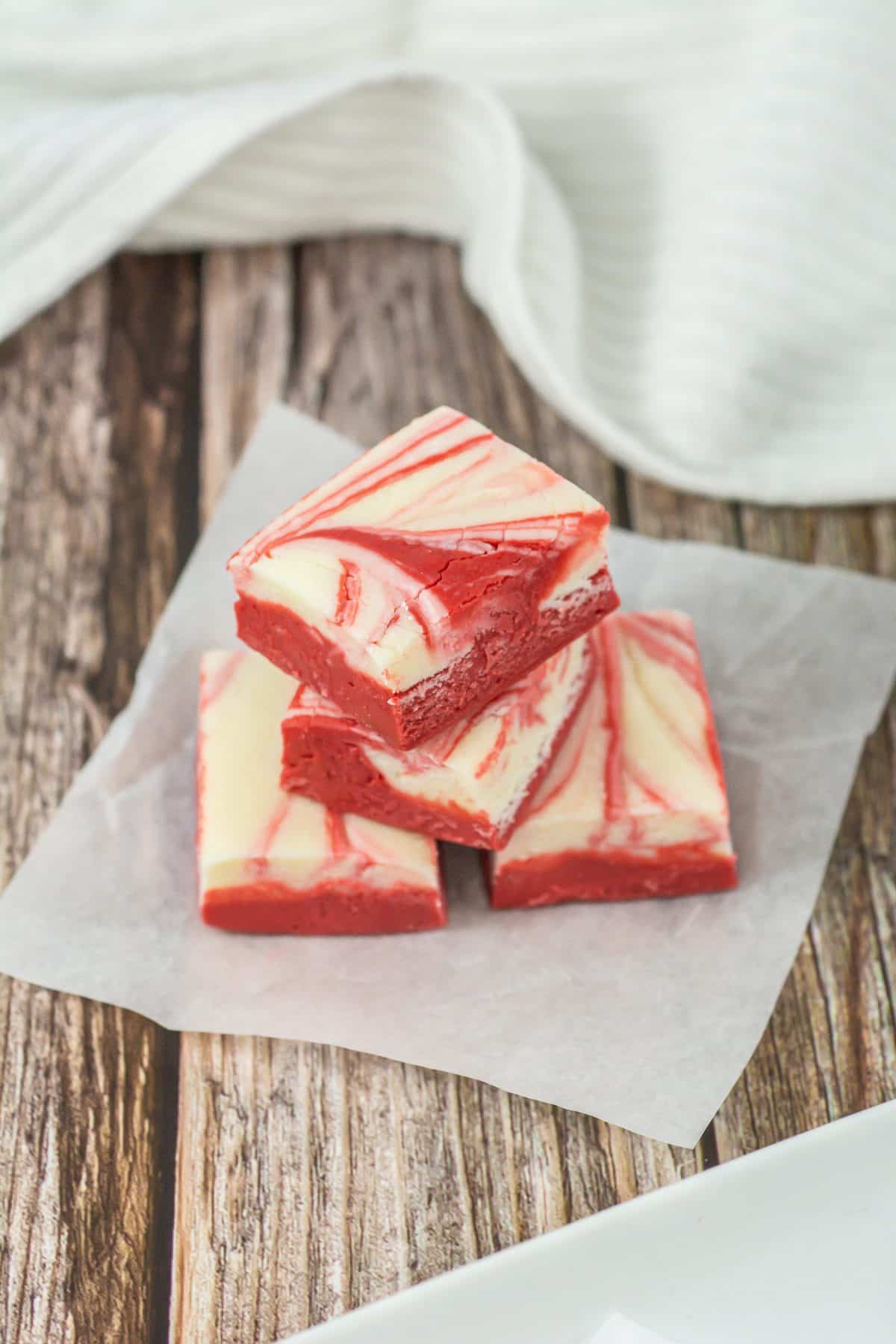 three squares of red velvet fudge stacked on a small square of white parchment paper on a wooden table