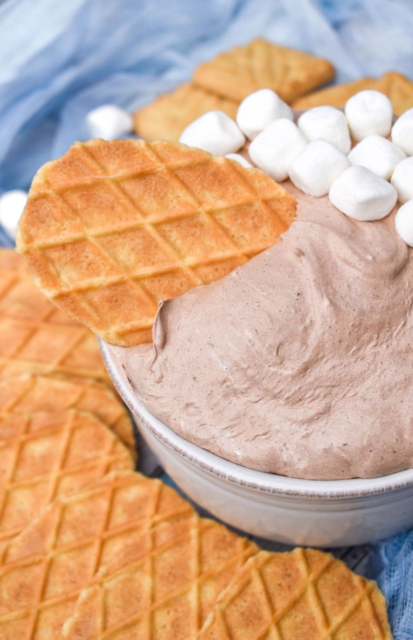 a waffle cone cookie stuck in a bowl of hot cocoa dip with marshmallows on the side