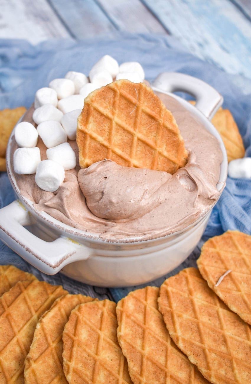 a waffle cone cookie stuck in a bowl of hot cocoa dip with marshmallows on the side