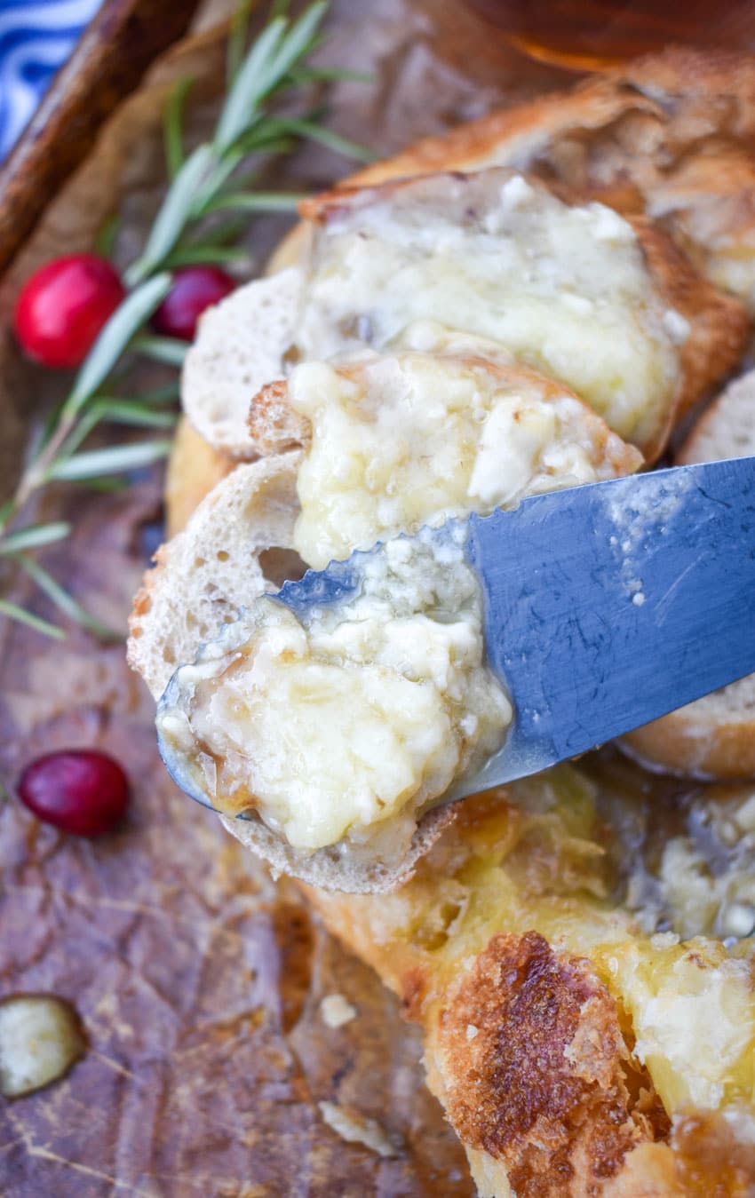 a cheese knife holding up a scoop of melted brie cheese over slices of baguette bread