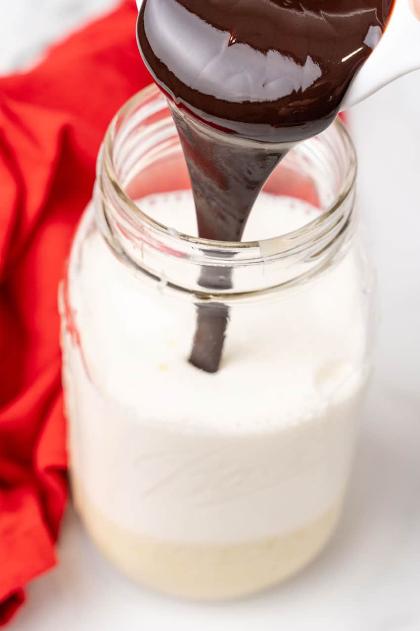 chocolate sauce being poured into a glass jar filled with sweetened condensed milk and half and half