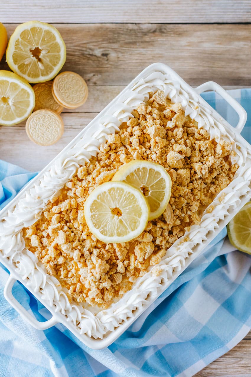 lemon oreo lasagna in a white casserole dish topped with crumbled oreo cookies and two slices of fresh lemon