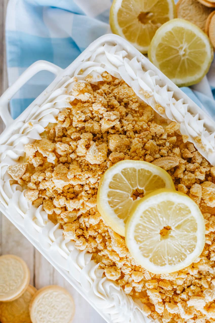 lemon oreo lasagna in a white casserole dish topped with crumbled oreo cookies and two slices of fresh lemon