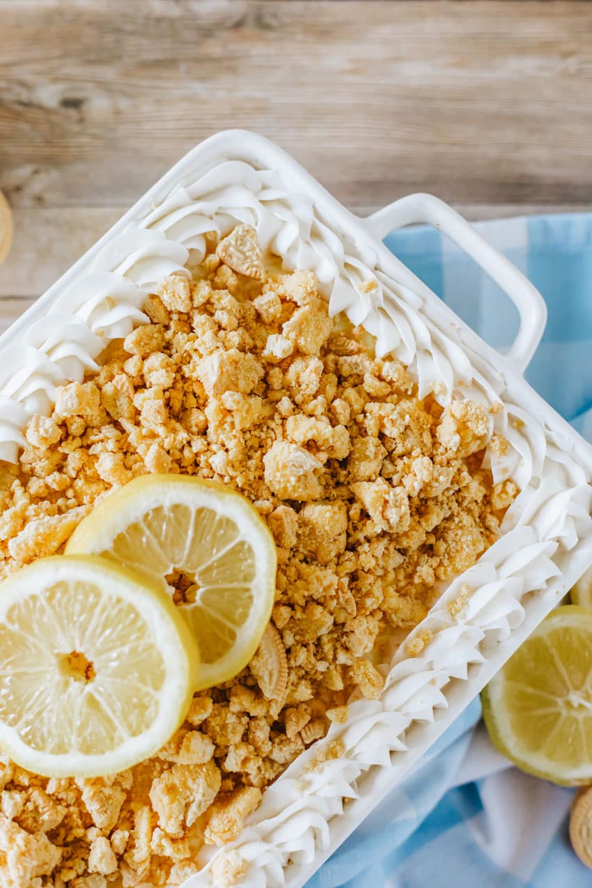 lemon oreo lasagna in a white casserole dish topped with crumbled oreo cookies and two slices of fresh lemon
