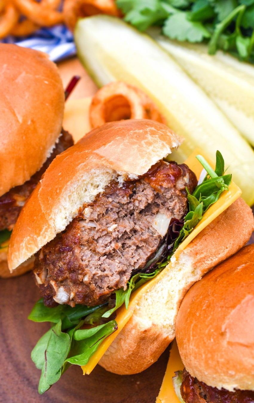 a bitten meatloaf burger on a wooden cutting board with dill pickle spears and crispy curly fries in the background