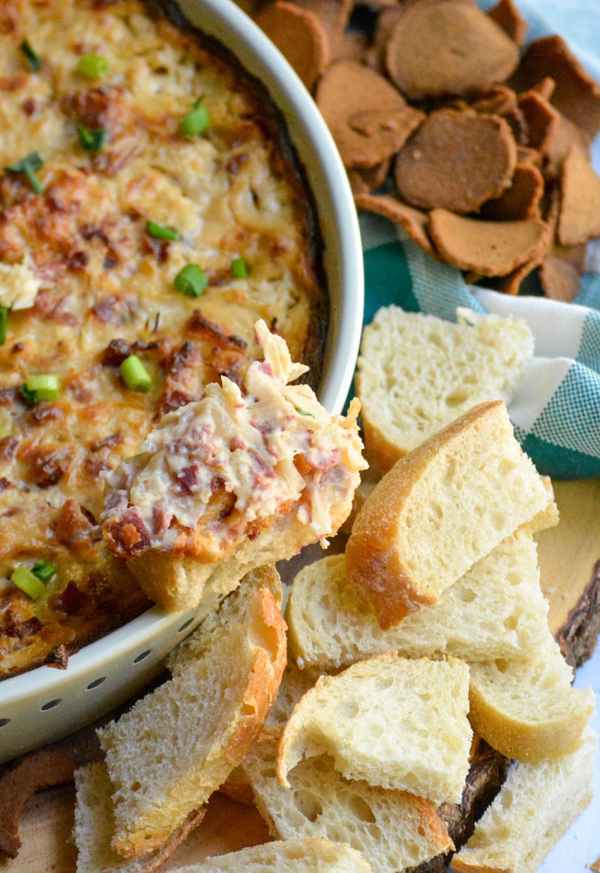 a baguette slice topped with baked reuben dip resting on the edge of a ceramic baking dish