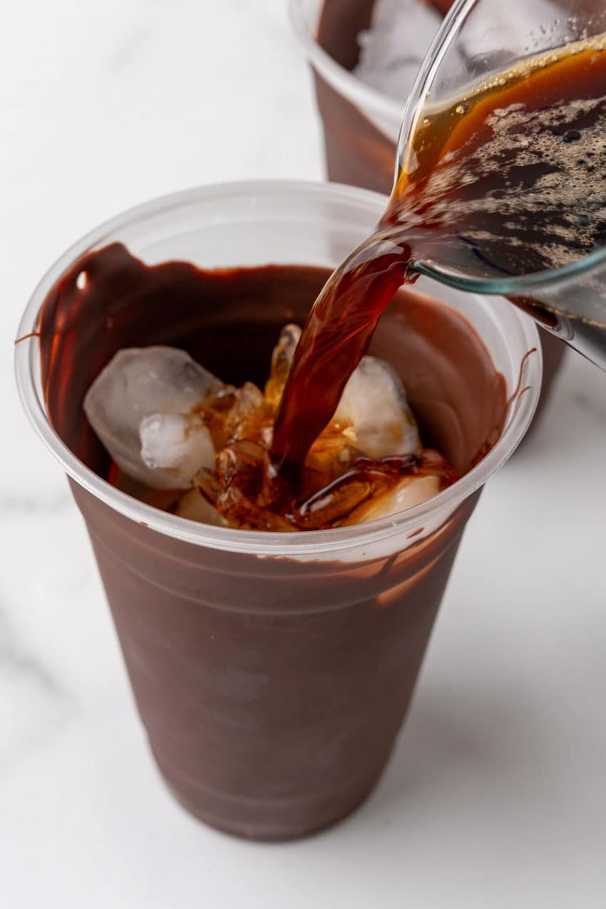 cold coffee being poured over ice into a chocolate coated plastic cup