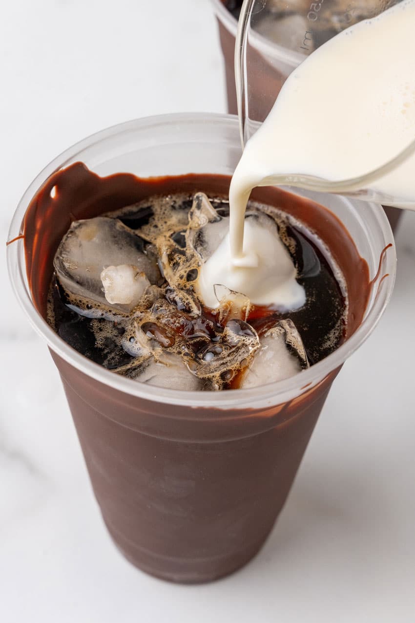 cream being poured over cold coffee over ice into a chocolate coated plastic cup