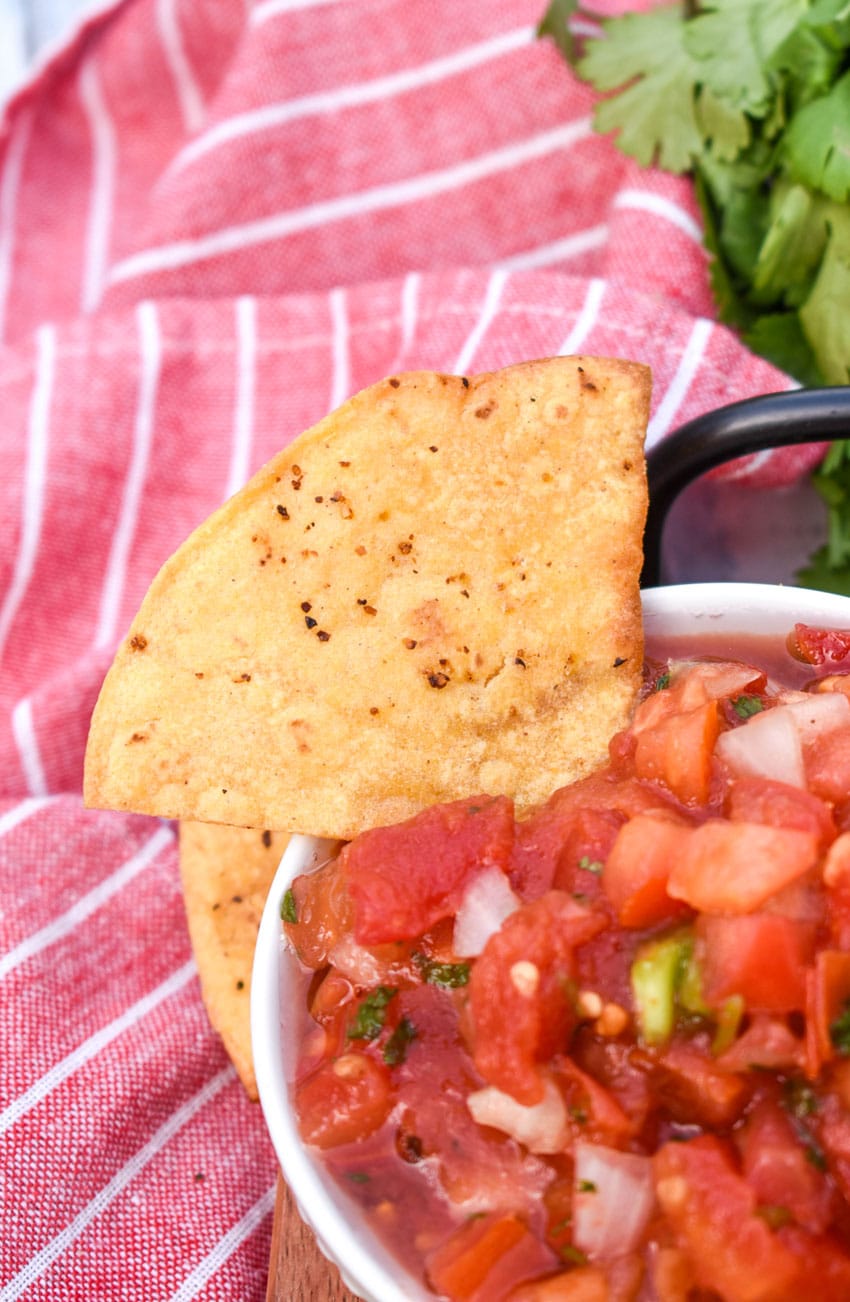 an air fried tortilla chip resting on the edge of a small white bowl filled with salsa