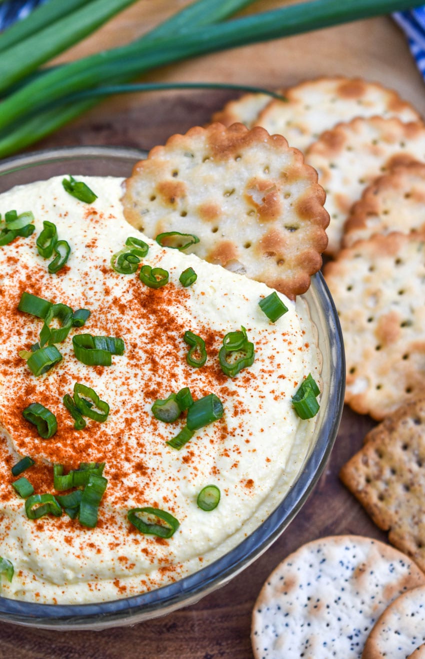 A CRACKER SCOOPING DEVILED EGG DIP OUT OF A SMALL GLASS BOWL