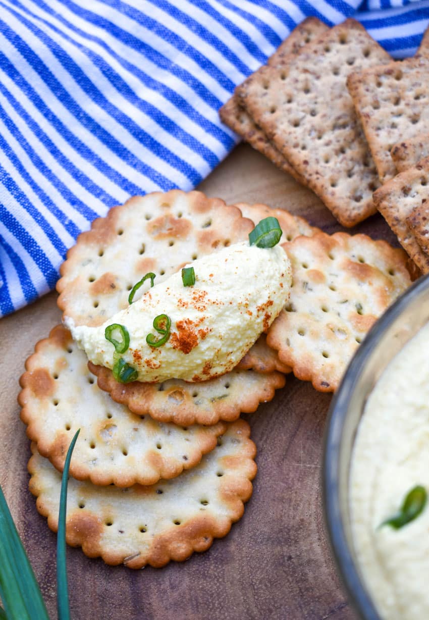 a cracker topped with deviled egg dip on a stack of crackers on a wooden cutting board