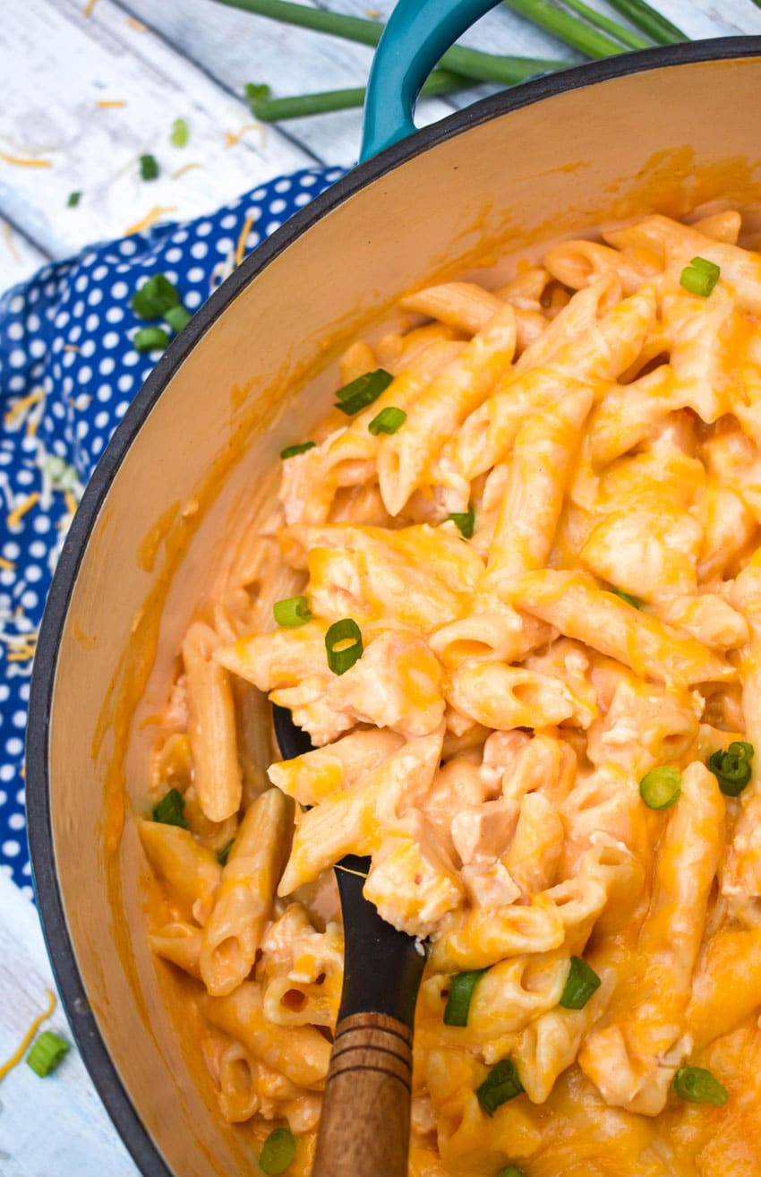 A WOODEN HANDLED SPOON SCOOPING CREAMY BUFFALO CHICKEN PASTA OUT OF A LARGE BLUE DUTCH OVEN