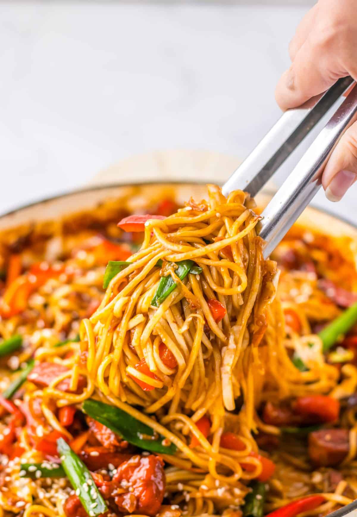 tongs lifting a scoop of pork lo mein out of a skillet