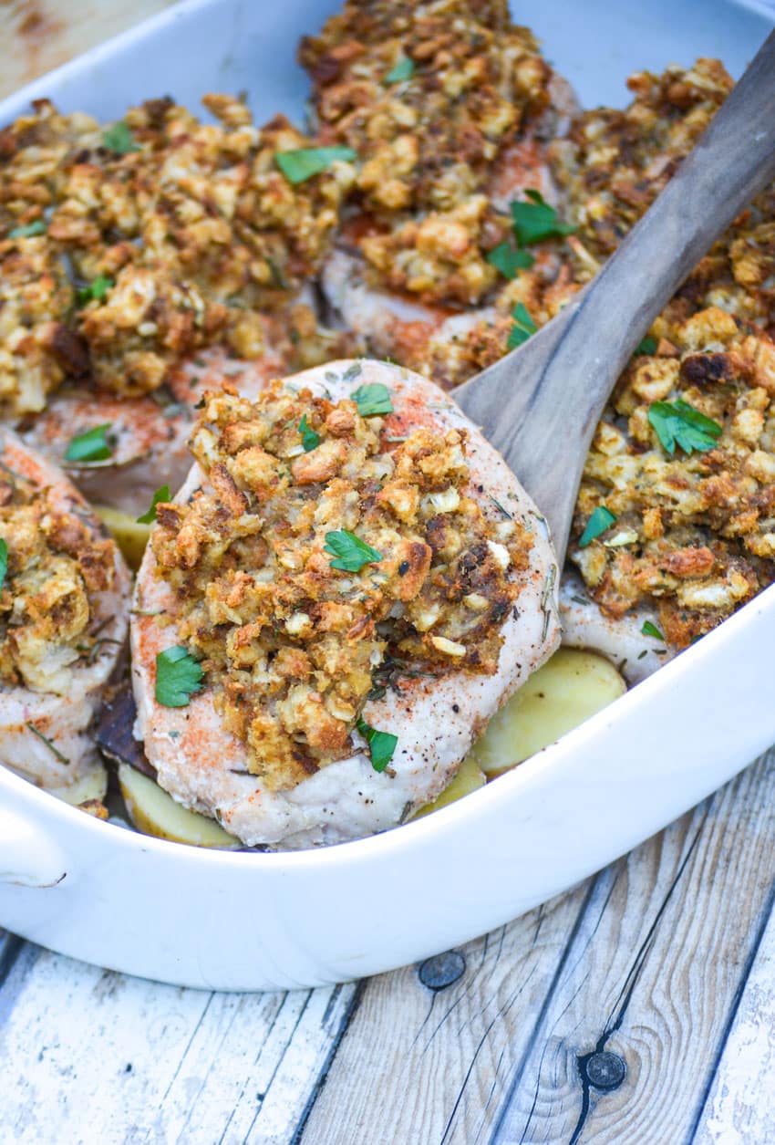 a wooden spoon scooping a stuffing topped baked pork chop out of a white baking dish