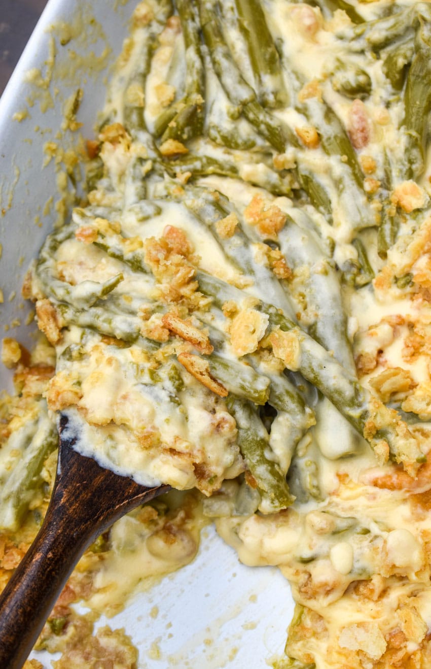 a wooden spoon scooping cheesy asparagus casserole out of a white baking dish