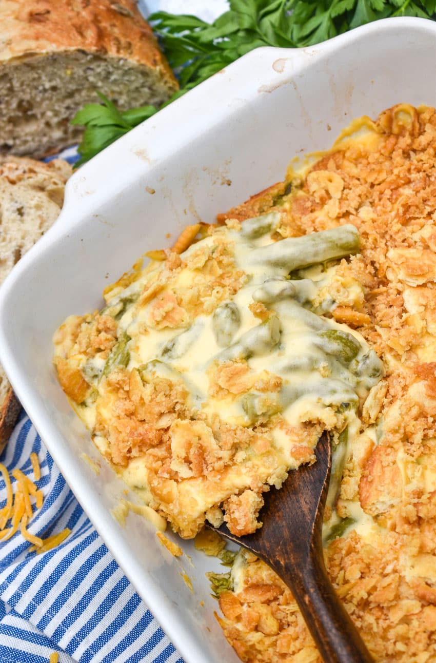 a wooden spoon scooping cheesy asparagus casserole out of a white baking dish