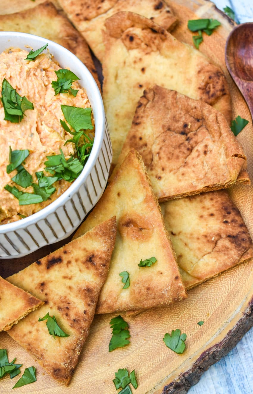 baked naan bread chips next to a small white bowl of chicken tikka masala dip on a wooden cutting board