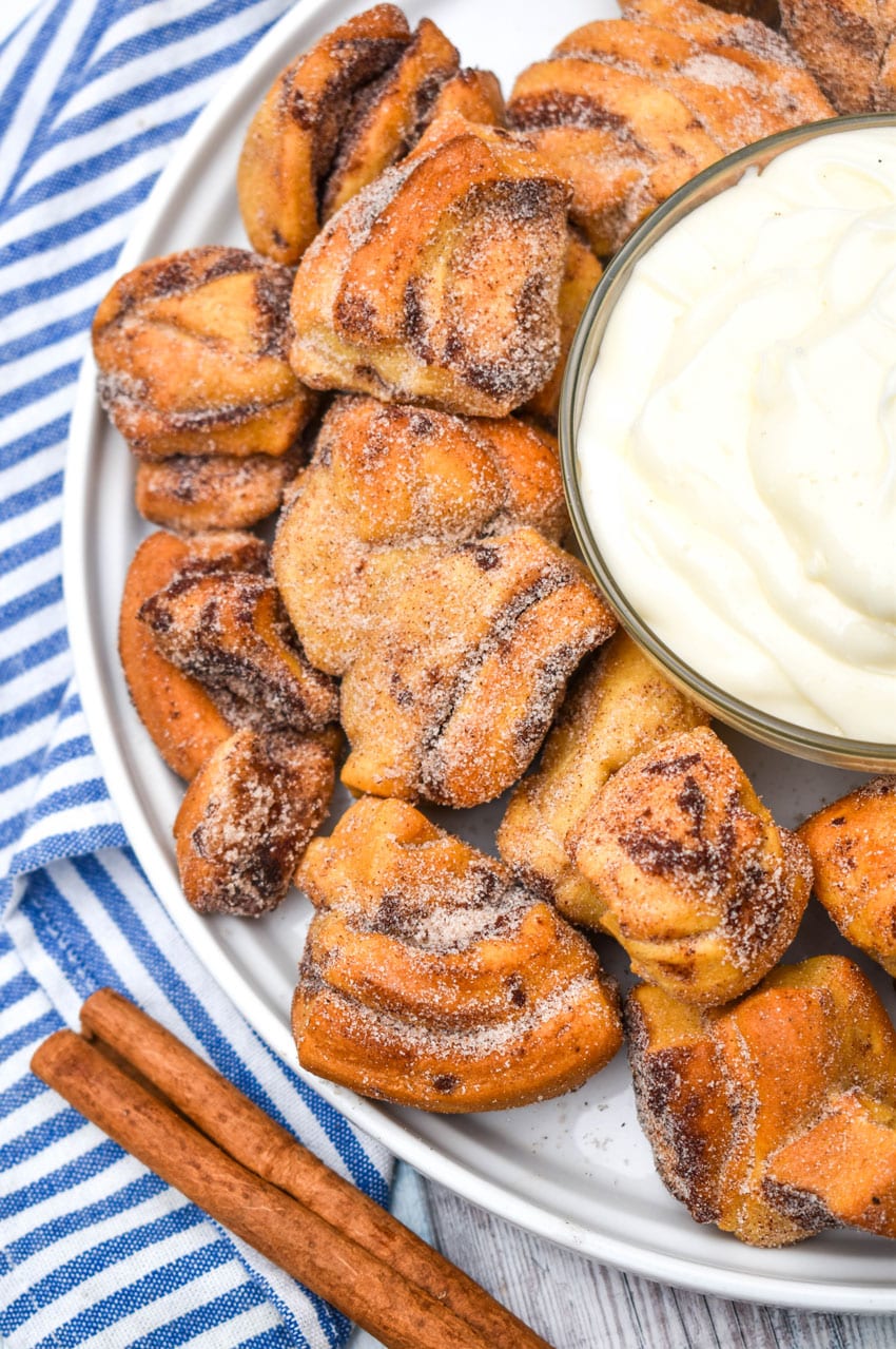 air fryer cinnamon roll bites surrounding a bowl of cream cheese icing on a white plate