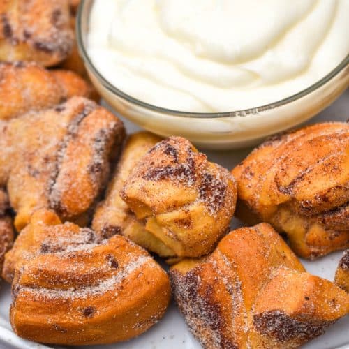air fryer cinnamon roll bites surrounding a bowl of cream cheese icing on a white plate
