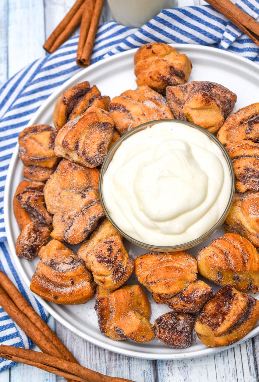 air fryer cinnamon roll bites surrounding a bowl of cream cheese icing on a white plate