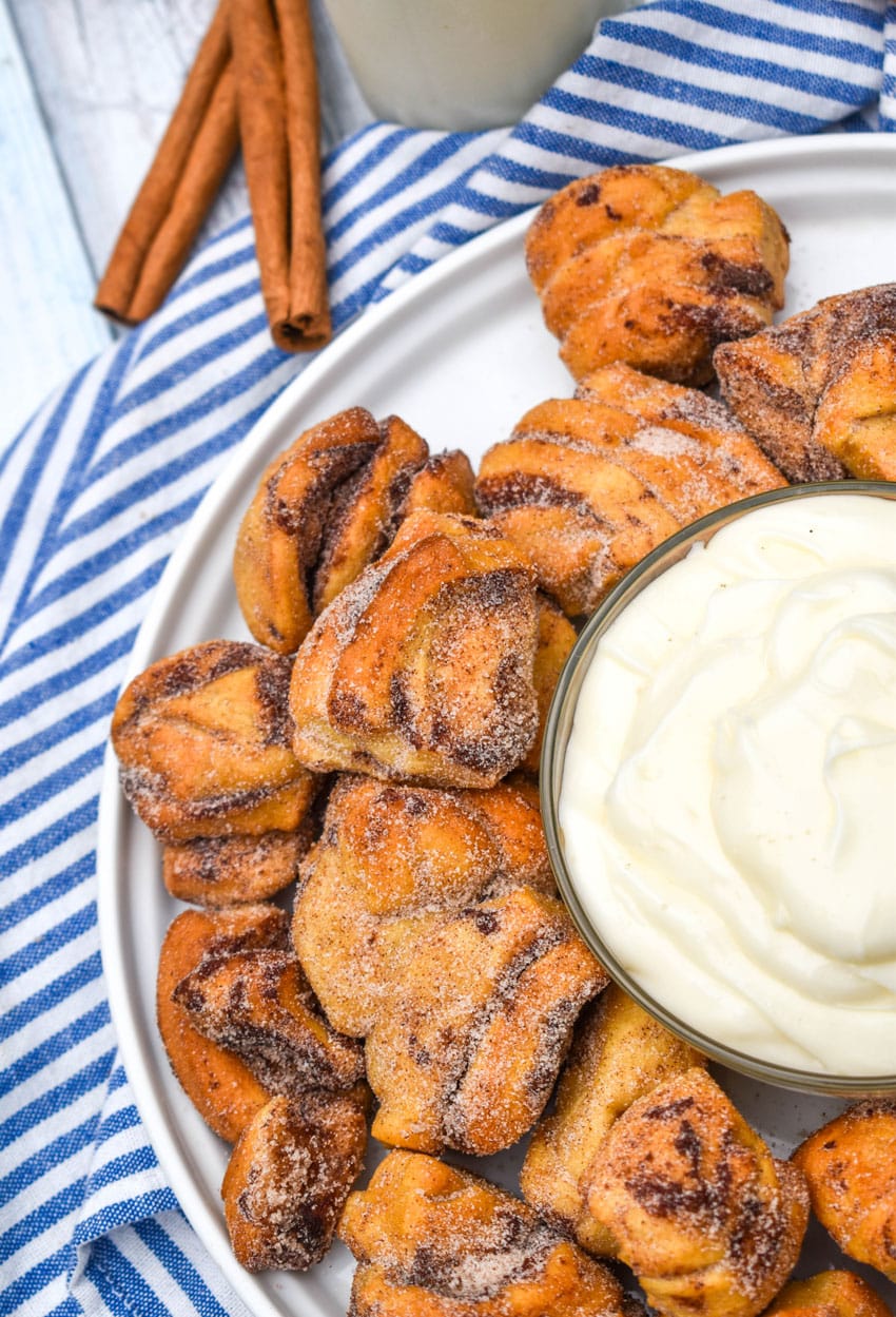air fryer cinnamon roll bites surrounding a bowl of cream cheese icing on a white plate