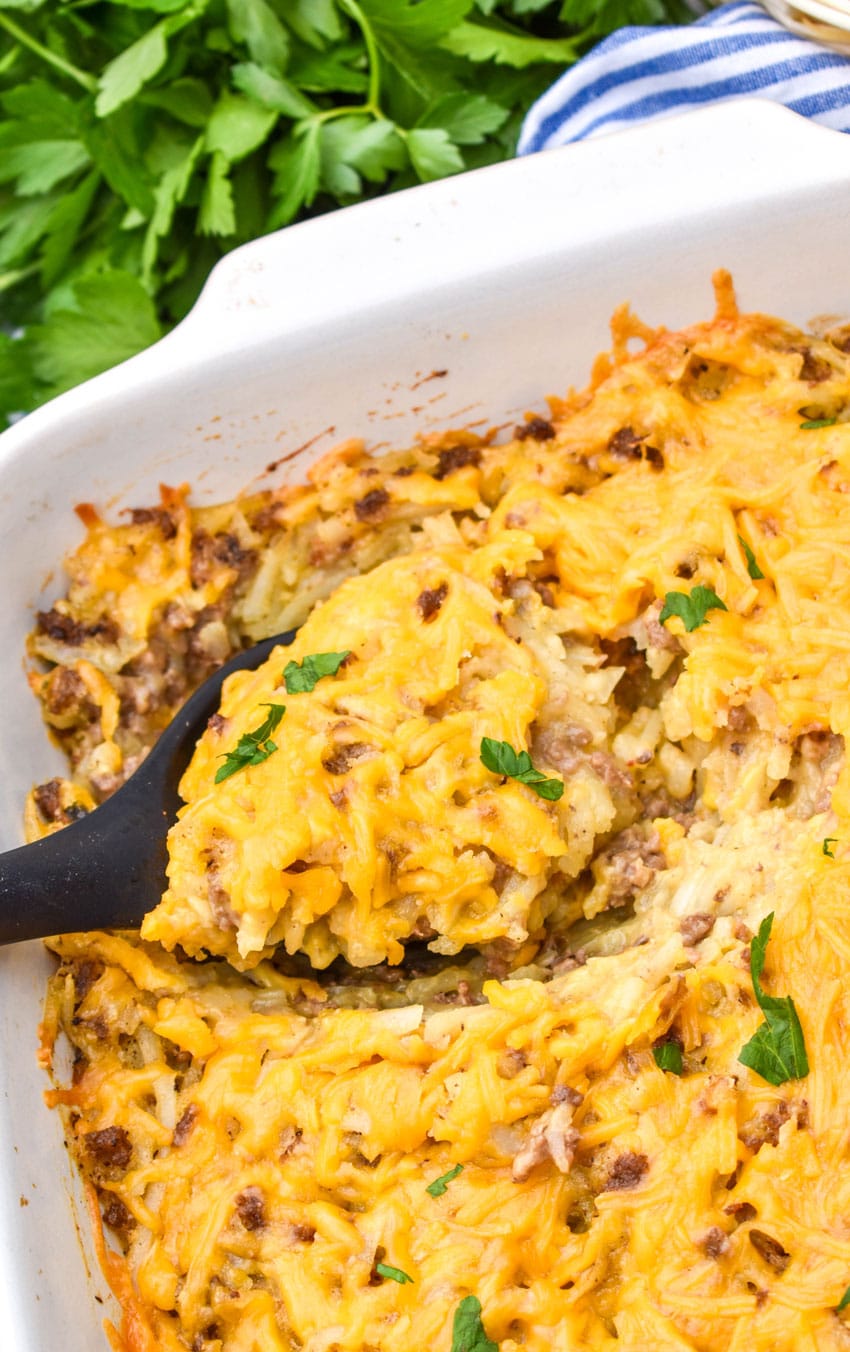 a black spoon scooping cheesy hashbrown casserole out of a white baking dish