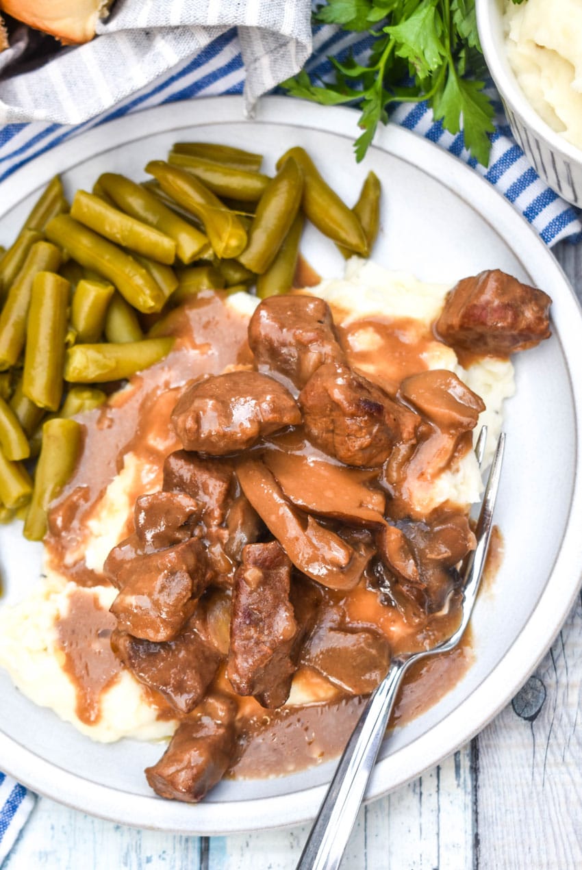 no peek beef tips over mashed potatoes next to green beans on a white dinner plate