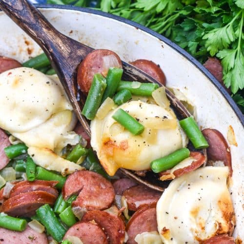 a wooden spoon scooping pierogi, kielbasa, and vegetables out of a cast iron skillet