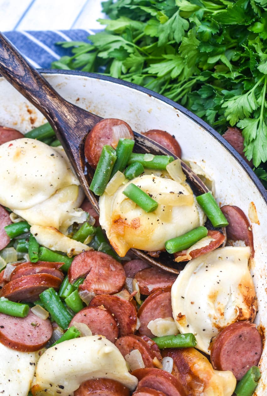 a wooden spoon scooping pierogi, kielbasa, and vegetables out of a cast iron skillet