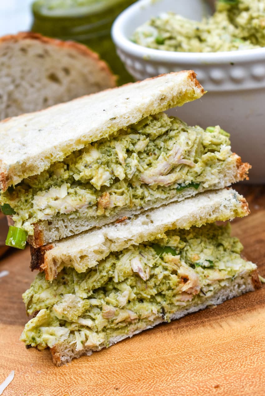 two halves of a chicken pesto salad sandwich stacked on a wooden cutting board