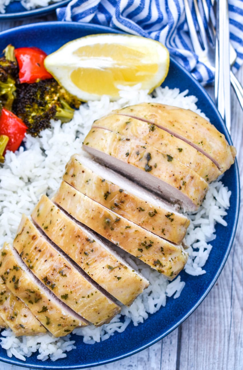 a sliced piece of sheet pan lemon chicken over steamed white rice on a blue plate with roasted vegetables on the side