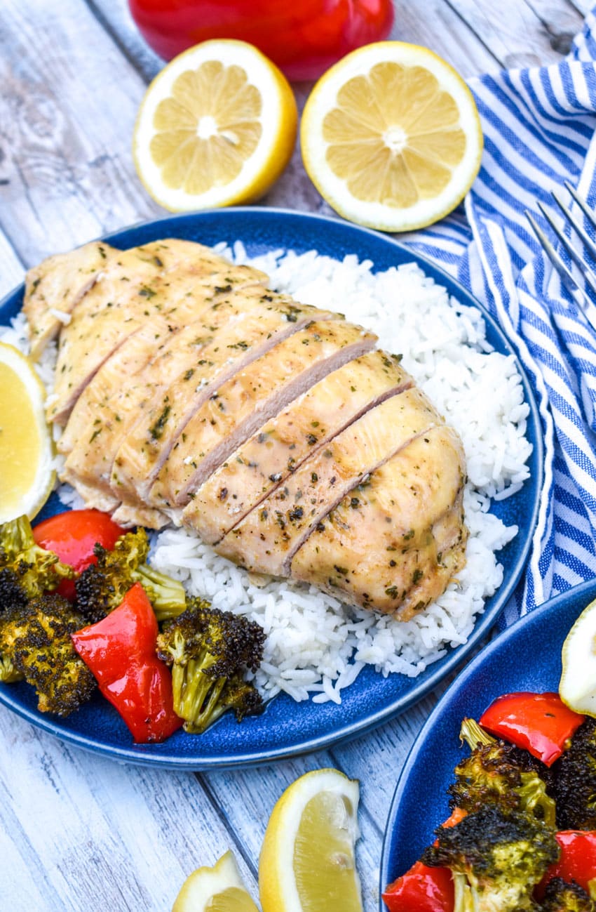 a sliced piece of sheet pan lemon chicken over steamed white rice on a blue plate with roasted vegetables on the side