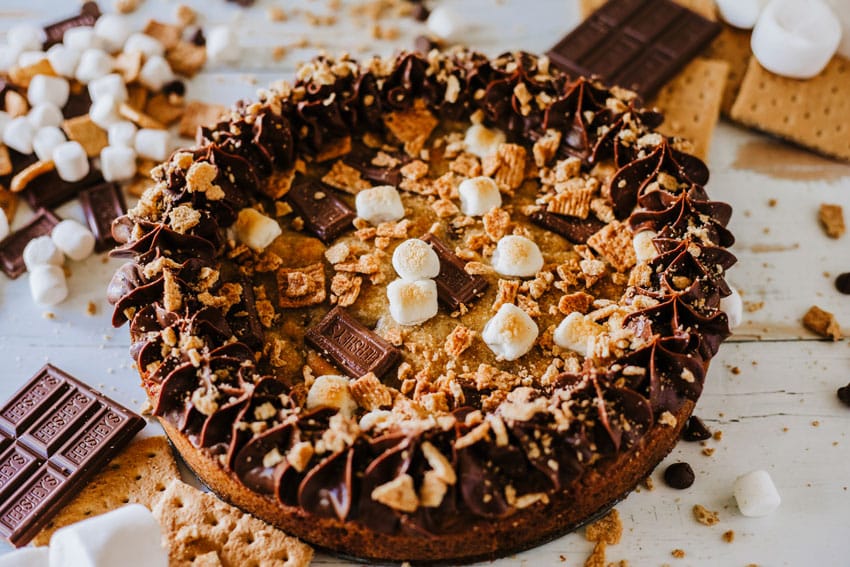a s'mores cookie cake on a wooden table surrounded by graham cracker crumbs and chocolate candy