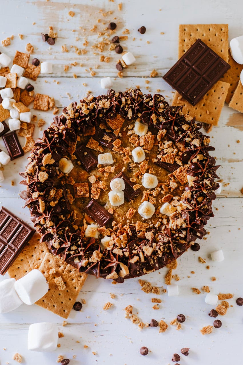 a s'mores cookie cake on a wooden table surrounded by graham cracker crumbs and chocolate candy