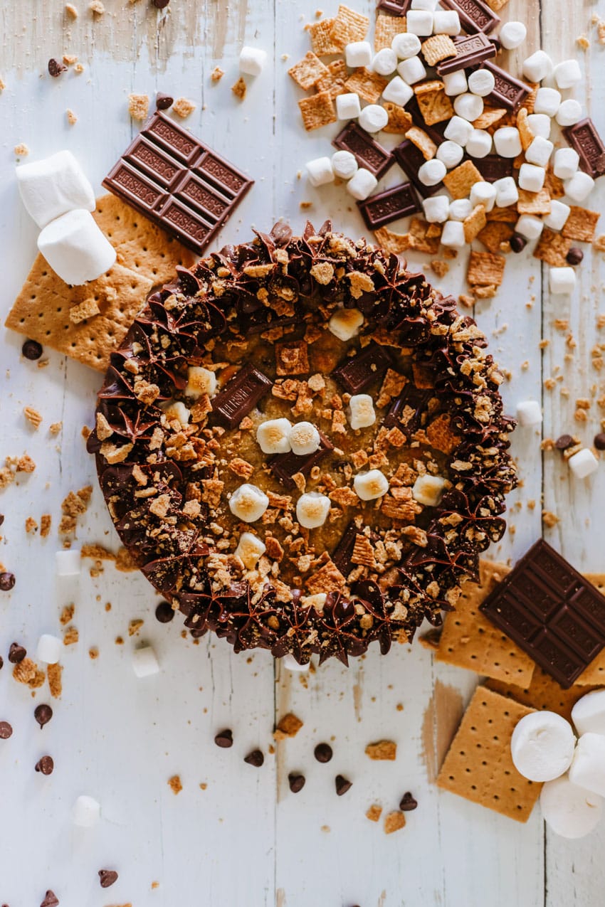 a s'mores cookie cake on a wooden table surrounded by graham cracker crumbs and chocolate candy