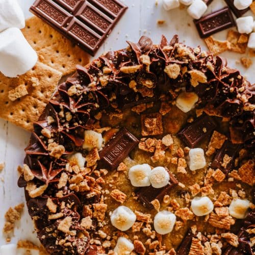 a s'mores cookie cake on a wooden table surrounded by graham cracker crumbs and chocolate candy