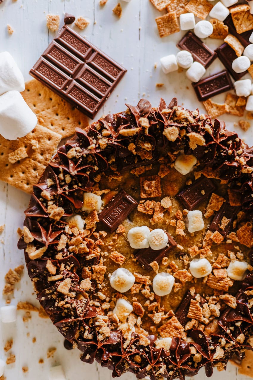 a s'mores cookie cake on a wooden table surrounded by graham cracker crumbs and chocolate candy