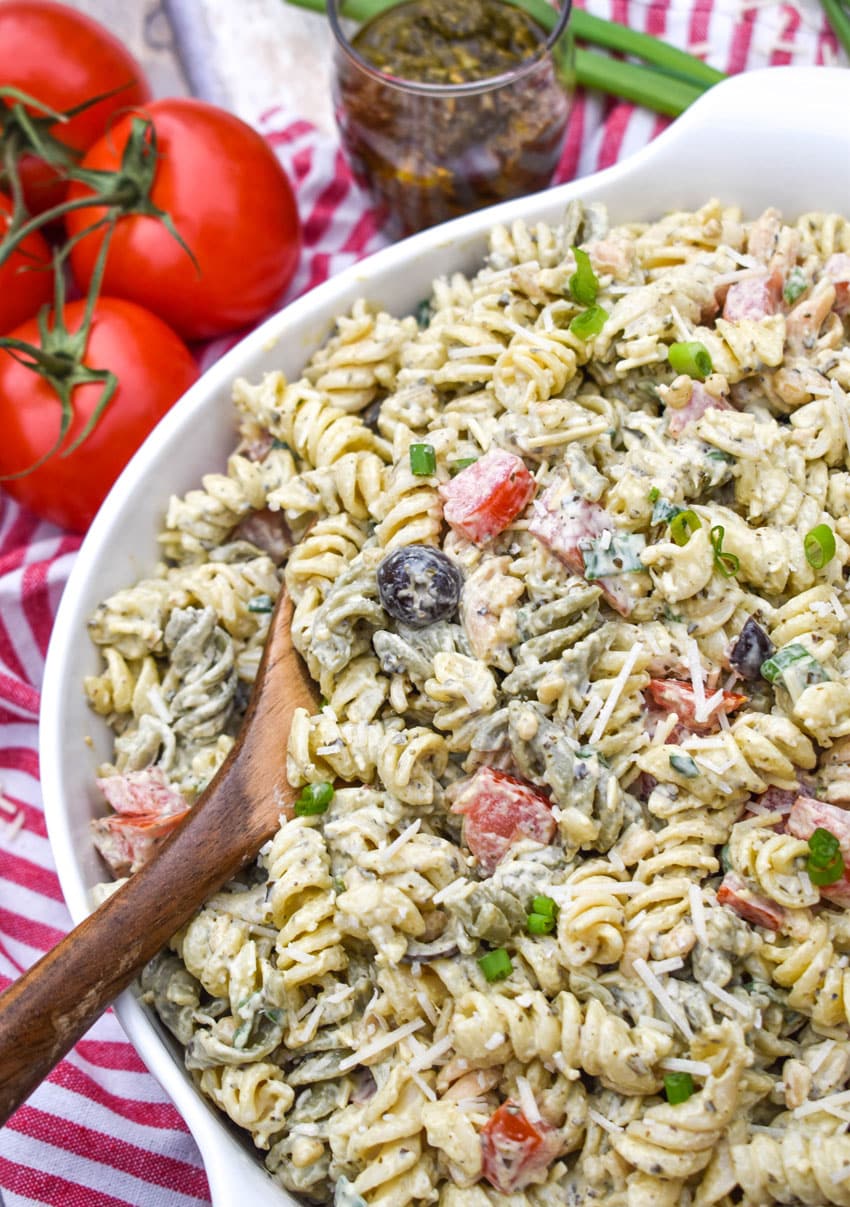 a wooden spoon scooping creamy caesar pesto pasta salad out of a white serving bowl
