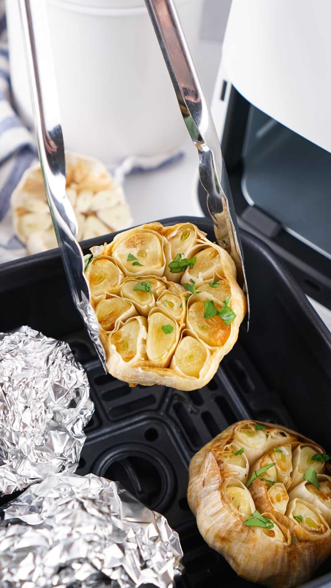 a pair of tongs lifted roasted garlic out of an air fryer basket